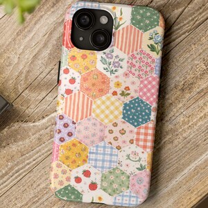 Könnte beinhalten: Eine bunte Handyhülle mit Patchwork-Design. Die Hülle zeigt eine Vielzahl von sechseckigen Mustern in Pastellfarben, darunter Streifen, Vichy-Karo, Blumenmuster und Erdbeermotive. Die Handyhülle liegt auf einer Holzoberfläche.