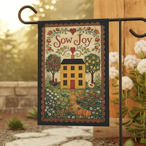 May include: A garden flag with a black border features a yellow house with a red door, flanked by fruit trees. The words "Sow Joy" are at the top, above the house. The flag is displayed on a black metal stand in a garden setting.