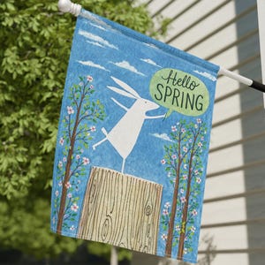 Peut inclure: Un drapeau bleu pour l'extérieur avec un lapin blanc sautant sur un bloc de bois, avec les mots "Hello SPRING" dans une bulle verte. Le drapeau a un fond de ciel bleu avec des nuages blancs et des arbres fleuris roses.