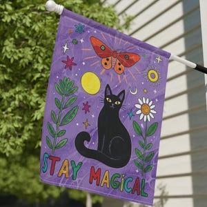 May include: A purple garden flag with a black cat, butterfly, and floral design. The flag has the words "STAY MAGICAL" in colorful letters. The flag is hanging outside on a white wall.