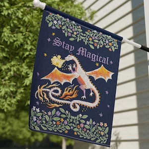 Puede incluir: Una bandera de jardín azul oscuro con un dragón blanco de estilo bordado que exhala fuego naranja y amarillo. El texto "Stay Magical" está impreso en una fuente decorativa sobre el dragón, rodeado de bordes florales y estrellas.