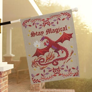May include: A rectangular garden flag with a light beige background featuring a red dragon breathing white smoke. The text "Stay Magical" is printed in red above the dragon. The design includes red and purple vine borders and yellow stars.