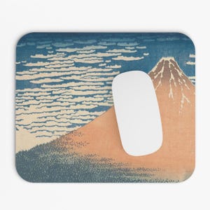 May include: A blue and white mouse pad with a Japanese woodblock print of Mount Fuji. The mountain is depicted in shades of pink, brown, and white, with a blue sky and white clouds in the background.