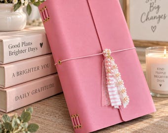 Könnte beinhalten: Ein rosafarbenes Notizbuch mit glattem Einband, das mit einem dünnen Gummiband verschlossen wird. Daran befindet sich ein dekorativer Anhänger aus weißen und rosa Perlen, kleinen Blumenakzenten und einem gestreiften Band. Goldfarbene Ösen.