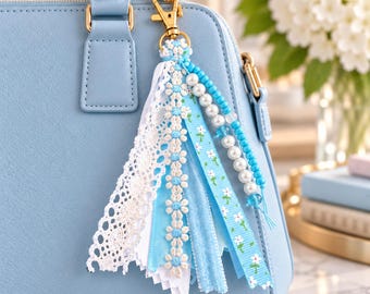 Könnte beinhalten: Ein hellblauer Handtaschenanhänger mit goldenem Karabinerverschluss, weißer Spitzenborte, einem blau-weißen Gänseblümchenband, einem hellblauen Band mit weißem Blumenmuster sowie einer Kette aus weißen Perlen und blauen Perlen.
