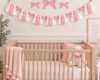Könnte beinhalten: Eine dekorative Girlande mit abwechselnden rosa und weißen Garnquasten, durchsetzt mit kleinen Holzperlen und rosa schleifenförmigen Ausschnitten. Die Girlande hängt horizontal und wird durch drei zusätzliche rosa Wandschleifen ergänzt.