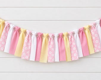 Könnte beinhalten: Eine Stoffgirlande mit einer Reihe geknoteter Streifen in abwechselnden Farben in Zartrosa, Blassgelb und Weiß. Einige der rosa Stoffstreifen sind mit einem kleinen weißen Blumenmuster versehen, die alle an einer zentralen Schnur hängen.