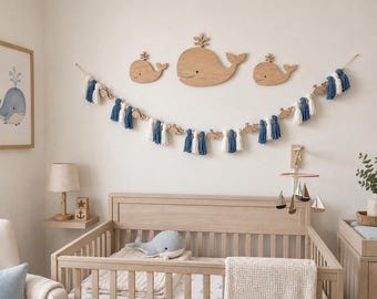 Könnte beinhalten: Ein Wanddekorationsset für das Kinderzimmer bestehend aus drei hellen Holzwalsilhouetten und einer langen Girlande mit abwechselnden dunkelblauen und weißen Stoffquasten, durchsetzt mit kleinen Walformen aus Holz und runden Perlen.