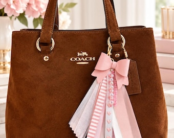 Könnte beinhalten: Eine braune Wildlederhandtasche mit zwei Tragegriffen, goldfarbenen Beschlägen und dem Coach-Logo in goldener Schrift. Am Griff hängt ein dekorativer Anhänger, bestehend aus einer rosa Schleife, einer Perlenkette und verschiedenen rosa und weißen Bändern.