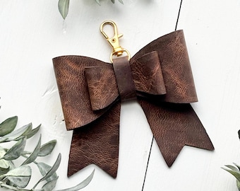 May include: A brown leather bow keychain with a gold-colored clasp. The bow has a textured appearance and is made of folded leather. The keychain is a decorative accessory.
