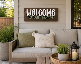 Könnte beinhalten: Ein rechteckiges Holzschild mit dunkelbrauner Oberfläche, das den weißen 3D-Schriftzug "WELCOME" und den grünen 3D-Schriftzug "to our porch" zeigt, montiert an der Wand über einem hellgrauen Sofa mit einem grünen Strukturkissen und einem cremefarbenen Kissen mit Quasten.