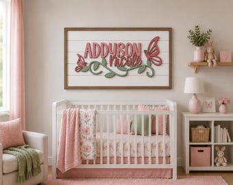 Könnte beinhalten: Ein rechteckiges Wandschild mit dem Namen Addyson Nicole in rosa Schreibschrift, verziert mit einer grünen Ranke und zwei rosa Schmetterlingen. Das Schild ist auf einem weißen Hintergrund in Holzoptik mit dunklem Rahmen über einem Kinderbett montiert.