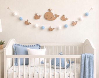 Könnte beinhalten: Eine Wandgirlande mit hölzernen Wal-Ausschnitten und abwechselnd weißen und hellblauen Pompons, die über einem weißen Kinderbett hängt. Das Bett enthält einen blauen Plüschwal, ein blaues Kissen, ein weißes Kissen mit Wal-Druck und eine blaue Decke.