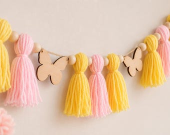 Könnte beinhalten: Eine dekorative Girlande mit abwechselnden pastellrosa und hellgelben Garnquasten, natürlichen Holzperlen und flachen Schmetterlingsausschnitten aus Holz, die auf einer dünnen Schnur aufgereiht sind und ein Muster aus weichen Textilelementen bilden.