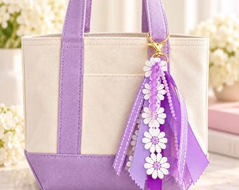 Könnte beinhalten: Eine cremefarbene Canvas-Tasche mit lavendelfarbenen Besätzen und Griffen. Am Griff befindet sich ein dekorativer Taschenanhänger mit weißen gänseblümchenförmigen Spitzenapplikationen, violetten Bändern und lila Perlen.