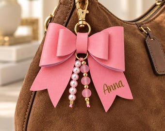 Könnte beinhalten: Eine braune Wildlederhandtasche mit einem rosa schleifenförmigen Taschenanhänger, der mit einem goldfarbenen Karabinerhaken befestigt ist. Der Anhänger hat zwei hängende Perlenstränge und den Namen Anna in goldener Schrift auf der rechten Seite.