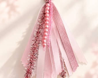 Könnte beinhalten: Ein dekorativer Quasten-Schlüsselanhänger mit einem goldfarbenen Karabinerhaken, bestehend aus einer Ansammlung von rosa Bändern, metallischen Lamettastreifen, einer Schnur aus hellrosa Perlen und einer Kordel aus kleinen, transparenten rosa Perlen.