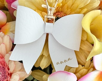 May include: A white leather bow keychain with a gold-colored clasp and the name "anna" in gold script. The keychain is surrounded by yellow, pink, and white flowers.
