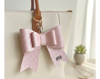 May include: A light pink leather bow bag charm with the name "ANNA" printed on one of the bow's tails. The bow is attached to a white handbag with a brown handle. A small succulent plant and a necklace are in the background.