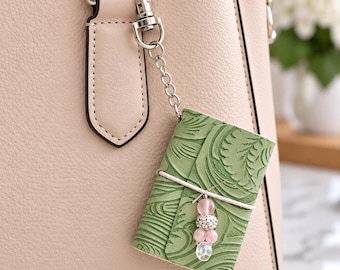 Könnte beinhalten: Eine hellbeige Handtasche ist mit einem kleinen, rechteckigen Anhänger aus salbeigrünem geprägtem Leder versehen, der an einer silbernen Metallkette befestigt ist. Der Anhänger wird von einem weißen Gummiband gehalten und mit rosa und silbernen Perlen verziert.