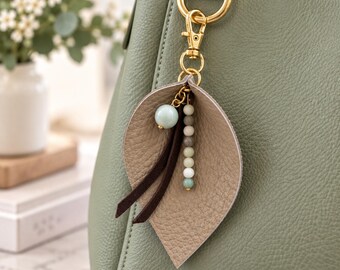 Könnte beinhalten: Eine salbeigrüne Handtasche mit einem dekorativen Taschenanhänger. Der Anhänger besteht aus einem goldfarbenen Karabinerverschluss, einer strukturierten beigen Lederblattform, zwei dunkelbraunen Wildlederstreifen und einer vertikalen Perlenkette.