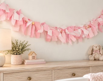 Könnte beinhalten: Eine Stoffgirlande aus miteinander verknoteten Stoffstreifen in Rosa, Weiß und Hellrosa. Die Girlande ist horizontal drapiert und enthält kleine Holzperlen, die zwischen den hängenden Stoffstücken eingearbeitet sind.