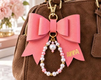 Könnte beinhalten: Ein Taschenanhänger aus rosa Kunstleder in Schleifenform mit goldenem Karabinerverschluss und einem tropfenförmigen Anhänger aus weißen und rosa Perlen mit goldenen Zwischenelementen. Auf einem Schleifenende steht der Name ANNA in Gold.