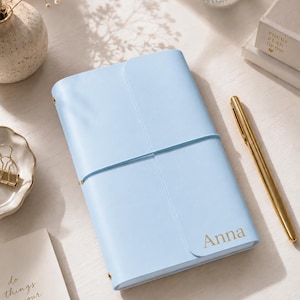 May include: A light blue rectangular notebook with a smooth cover and a vertical elastic band closure. The name Anna is printed in gold lettering on the bottom right corner of the cover. The notebook is positioned next to a gold pen.