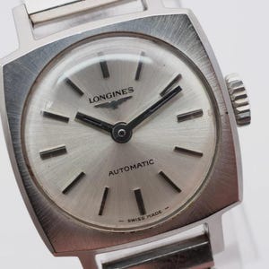 Könnte beinhalten: Eine Vintage Longines Automatik-Armbanduhr mit silbernem Zifferblatt und Edelstahlgehäuse und -armband. Das Zifferblatt der Uhr hat schwarze Zeiger und Markierungen sowie die Wörter "LONGINES" und "AUTOMATIC". Die Uhr ist mit "SWISS MADE" gekennzeichnet.