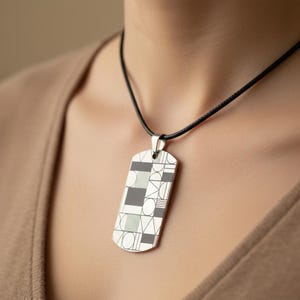 May include: A silver-toned rectangular dog tag pendant suspended from a thin black cord. The pendant features a geometric pattern consisting of black and white squares, circles, triangles, and vertical line segments arranged in a grid-like layout.