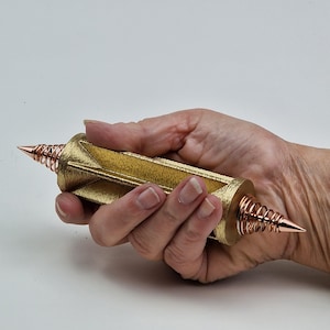 Può includere: Un oggetto geometrico dorato con punte coniche e spirali color rame. L'oggetto è tenuto in mano su sfondo bianco. Il corpo centrale ha una superficie strutturata.