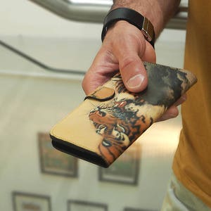 K&ouml;nnte beinhalten: Eine braune Ledergeldb&ouml;rse mit Tiger-Druck. Die Geldb&ouml;rse ist geschlossen und hat eine Klappe mit einem Knopfverschluss.