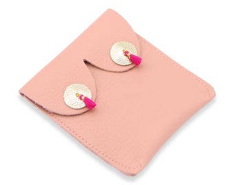Könnte beinhalten: Eine rosafarbene Leder-Pochette mit Klappenverschluss. Die Pochette hat zwei goldfarbene runde Akzente, die jeweils mit einer kleinen, leuchtend rosa Quaste verziert sind. Das Leder hat eine strukturierte Optik und die Pochette ist zum Falten konzipiert.