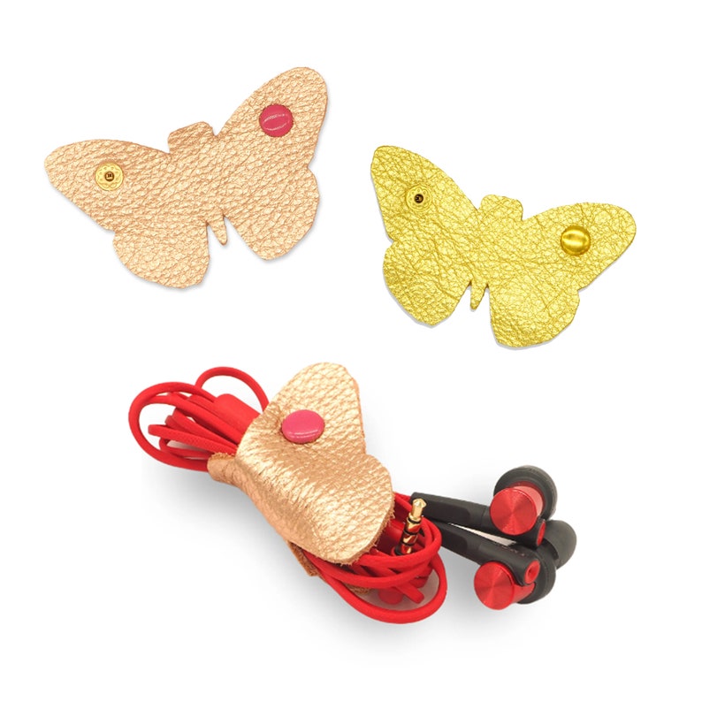 May include: Three gold leather butterfly shaped cord organizers with pink and gold buttons. One organizer is holding a pair of red earbuds.