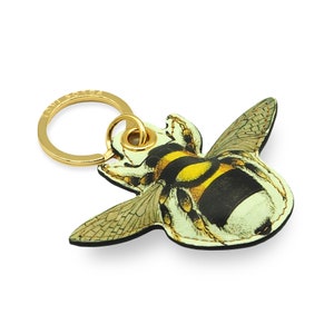 Puede incluir: Un llavero dorado con un dije de abejorro de cuero. La abeja es negra, amarilla y blanca con alas detalladas.