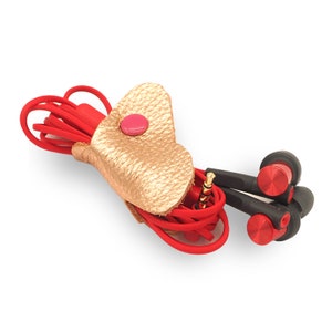 May include: A red cord with a gold leather heart-shaped cord organizer. The organizer has a pink button closure. The cord is connected to a pair of black and red earbuds.