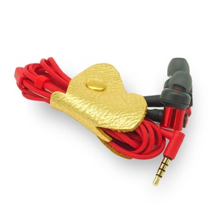 May include: A gold leather cord keeper with a red cord and black earbuds. The cord keeper has a gold button closure.