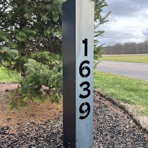 Peut inclure: Un poteau d'adresse rectangulaire en métal gris avec le numéro "1639" en noir. Le poteau est planté dans un lit de paillis sombre, avec un arbre vert et un ciel nuageux en arrière-plan.