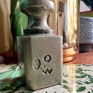 Könnte beinhalten: Ein verwittertes, silberfarbenes Metallgewicht mit quadratischer Basis und abgerundeter Oberseite. Die Vorderseite des Gewichts zeigt ein eingraviertes Gesicht mit einem kreisförmigen Auge, einem gebogenen Mund und einem Symbol. Das Gewicht liegt auf einer gemusterten Oberfläche.