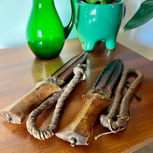 May include: Two antique daggers with wooden handles and leather-wrapped grips rest on a wooden surface. The daggers have a dark, aged appearance. A green glass pitcher and a green ceramic planter with a plant are in the background.