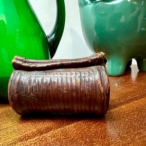 Puede incluir: Un pequeño recipiente cilíndrico antiguo hecho de cuero marrón oscuro texturizado. El recipiente tiene una parte superior redondeada y está decorado con líneas verticales y pequeñas hendiduras circulares. El fondo incluye objetos de vidrio verde.