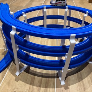 Könnte beinhalten: Ein blaues und weißes Spielzeug-Eisenbahn-Gleis-Set mit drei Ebenen kreisförmiger Gleise. Die Gleise sind aus Kunststoff und für die Verwendung mit Spielzeugeisenbahnen konzipiert. Die Gleise sind spiralförmig angeordnet, wobei die oberste Ebene der Gleise die höchste ist. Die Gleise werden von weißen Kunststoffstützen getragen.