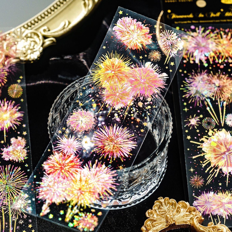 May include: Decorative washi tape featuring a fireworks design in shades of pink, yellow, and gold. The tape is transparent with a black background and is partially wrapped around a clear glass bowl. The design includes stars.