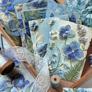 May include: A collection of rectangular cards featuring raised, textured blue and white floral relief designs. Each card displays detailed botanical elements including pressed-style flowers, ferns, and leaves in shades of blue, cream, and green.