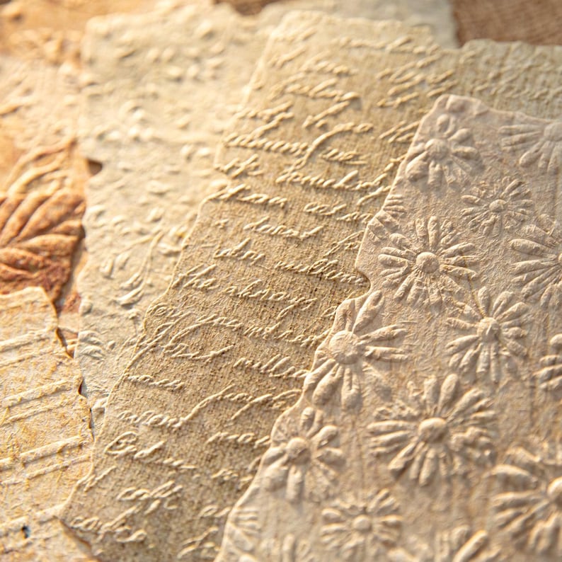 May include: Close-up of several textured, aged paper sheets. The paper has embossed floral and script designs in a warm, neutral color palette. The paper appears to be handmade, with a vintage aesthetic.