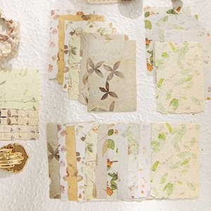 May include: A collection of handmade paper sheets with botanical designs. The papers feature pressed leaves, flowers, and other natural elements in shades of green, brown, and cream. Some sheets have deckled edges, adding a rustic touch.