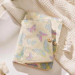 May include: Cream-colored journal with a butterfly pattern in pastel blue, pink, and yellow. The journal has a textured fabric cover and is secured with wooden button closures. The design features various sizes of butterflies.