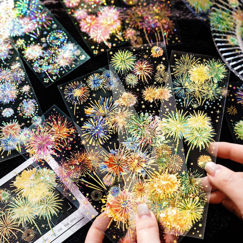 May include: Clear sticker sheets featuring colorful fireworks designs in shades of gold, orange, blue, and green. The stickers have a holographic effect, creating a shimmering appearance. The sheets are held by a person's hand.