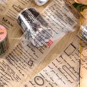 Op de afbeelding: Decoratieve washi tape rollen met vintage ansichtkaart ontwerpen. De tape heeft handgeschreven tekst, postzegels en decoratieve randen. De transparante tape is ideaal voor scrapbooking, journaling en knutselprojecten.