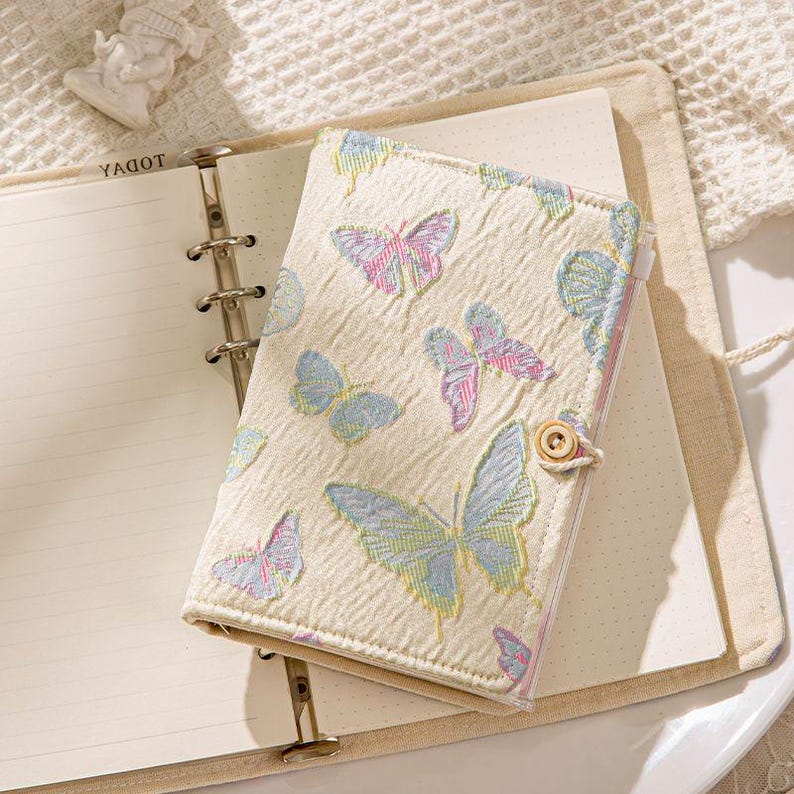 May include: Cream-colored journal with a butterfly pattern in pastel pink, blue, and yellow. The journal has a button closure and is open to reveal lined pages. The word "YAGOT" is printed on the top left page.